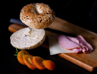 Deconstructed multigrain bagel sandwich with flying sliced pieces, tomato, ham and cheese placed on wooden cutting board with a knife on top of it.