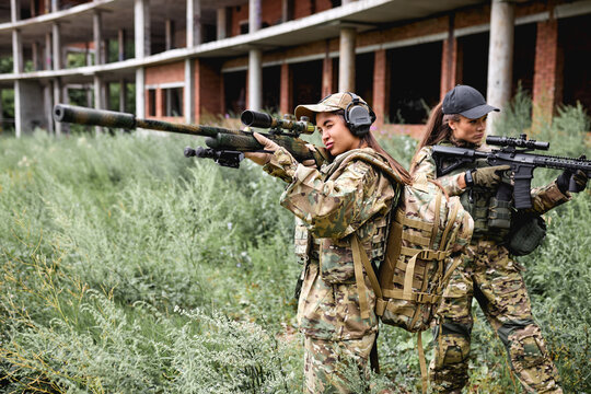 Confident army ladies women in military gear posing for photo in field at summer season. Female in green camo uniform and rifle with suppressor aiming at target, abandoned building in the background
