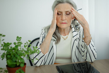 Tired senior grey haired businesswoman in striped jacket with eyeglasses is working in her office...