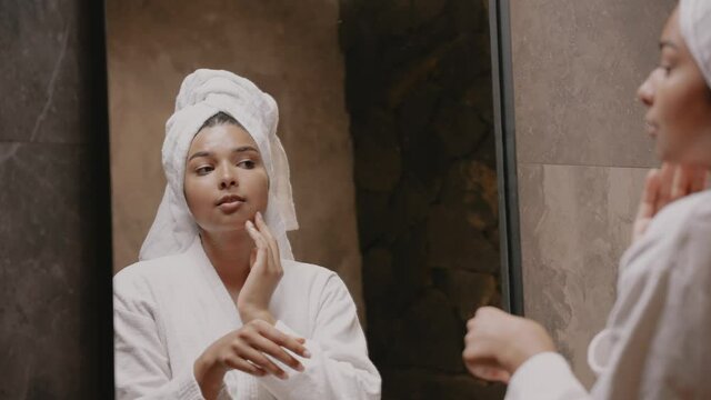 Young dark-skinned brunette in front of a mirror in a dressing gown and with a towel on her head applies cream to her face, the girl is reflected in mirror. Morning routine. High quality 4k footage
