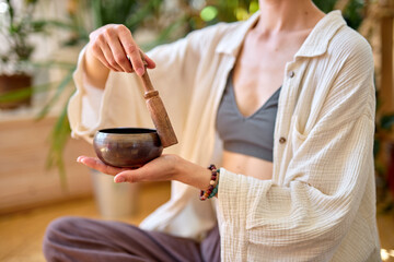 Woman using singing bowl in sound healing therapy at home, sitting alone at home, caucasian female in domestic wear pajamas begins morning with yoga practice, sit relaxed. Close-up photo of hands