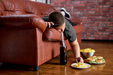 Fat Lazy Boy Taking Fries From Plate Lying On Sofa, Alone At Home, Teenager Boy Having Rest After School, Relaxed. Caucasian child lead unhealthy lifestyle, eating junk meal. overweight, obesity - obrazy, fototapety, plakaty