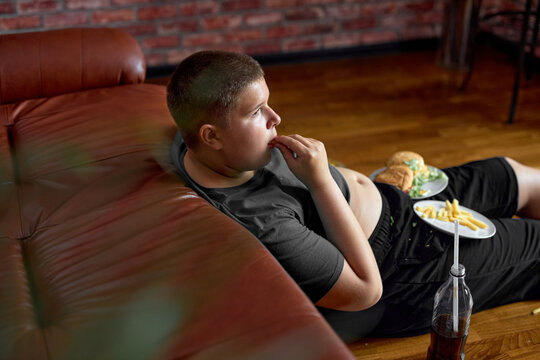 hungry caucasian teenager boy is eating Unhealthy junk fast food and watching tv sitting on floor. alone in dark living room. bad nutrition, diet, overweight, obesity concept