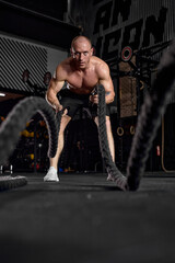 Fototapeta na wymiar Waves of force. Young caucasian healthy bodybuilder training with battle ropes. Fit strong single male model training with battle rope. Concept of sport, bodybuilding, healthy lifestyle.