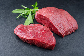 Raw dry aged bison beef rump steak piece and slices with herbs offered as close-up on black background