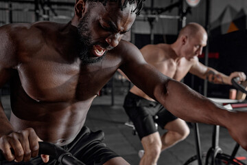 Fototapeta na wymiar Two diverse sportsmen riding stationary bike while working out at gym, cross fit cardio training, shirtless black and caucasian men have intense training. sport, healthy lifestyle. focus on afro male