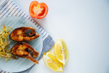 Fried fish with onions lies on a plate, on a waffle towel, next to a lemon and a tomato on a white background.