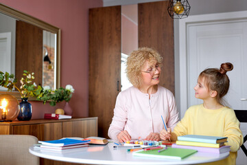 Pretty caucasian grandchild and senior gray-haired grandmother sitting at home drawing, doing homework. Smiling cute girl and senior woman use paints. side view portrait