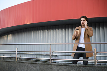 Businessman in coat talking on cellphone and holding coffee near building on urban street 