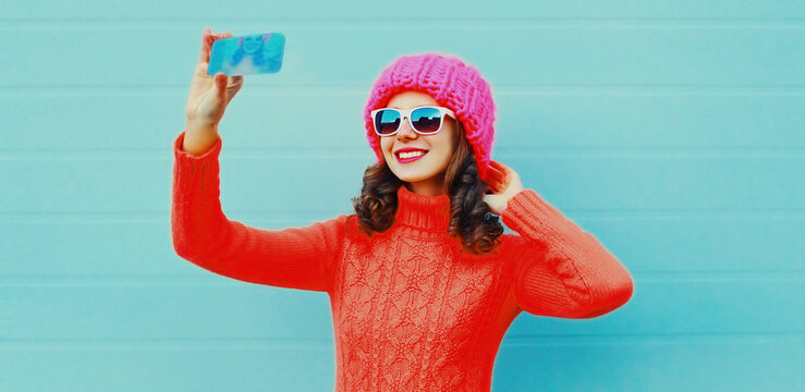 Portrait of happy smiling young woman taking a selfie by phone wearing a pink knitted hat, sweater on blue background