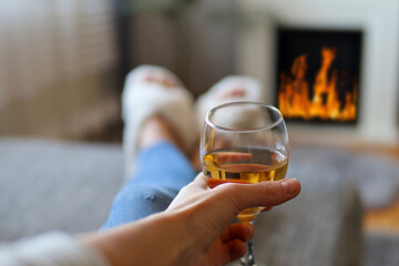 A woman lies in bed and holds glass of white wine and warms her legs on the fireplace