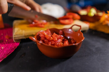Chopped tomato in small clay pot with hands slicing more tomato as background