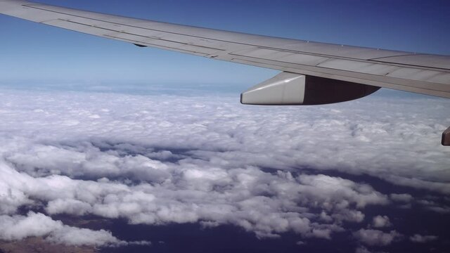 Travel flying above puffy clouds, looking to the left of the frame. Passenger window view of unbranded wing over blue horizon, aboard an airplane cruising at high altitude.