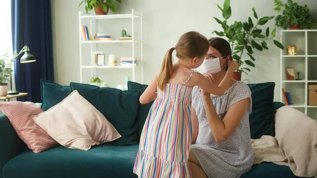 Little daughter putting on medical protective mask for her mother, taking care of parents health. Woman and child posing in living room at home. Coronavirus lockdown isolation concept.