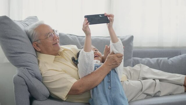 Happy senior grandfather with grandson taking photo by smartphone, old grandpa embracing little grand child take selfie on mobile phone together sit on sofa in living room at home, elderly and kid