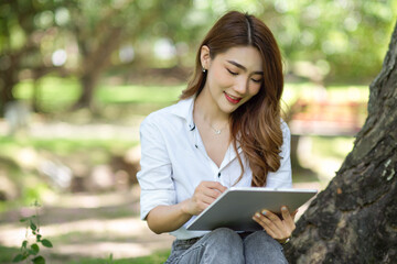 Fototapeta premium Beautiful Asian female sits in the park with a digital tablet