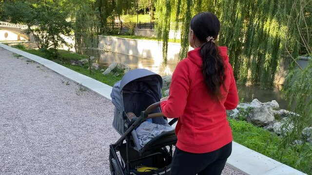 Mother with baby stroller on a walk outdoors, child sitting in gray bassinet, mom with six month old baby boy in infant kid carriage in park on a sunny summer day. Simferopol, Crimea, Russia