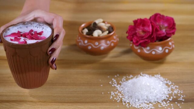 A lady serving a glass of chilled Chaas decorated with rose petals. Cashew nuts  raisins  and white sugar particles beautifully placed on a table - low in sugar  healthy heart  plant protein