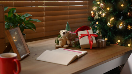 Notebook, gift boxes and teddy bear on wooden table near the Christmas tree in living room.