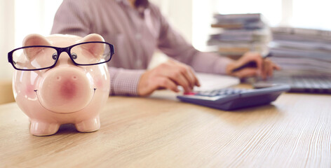 Banner with close up of cute piggy bank wearing eyeglasses placed on table of accountant who's...