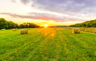 Scenic view at beautiful sunset in green shiny field with hay stacks, bright cloudy sky, country road and golden sun rays with glow, summer valley landscape