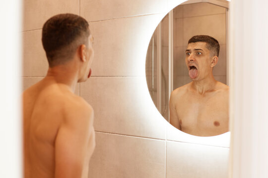 Indoor shot of young adult brunette male with naked upper body showing tongue in front of mirror in bathroom, doing morning hygienic procedures in the morning.