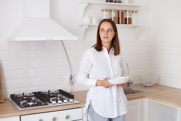 Indoor shot of attractive pensive dark haired young adult woman having breakfast in the kitchen, standing with plate in hands, looking away with thoughtful expression.