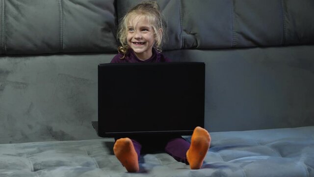 A girl child sits with a laptop on a gray couch and smiles. The concept of digital childhood