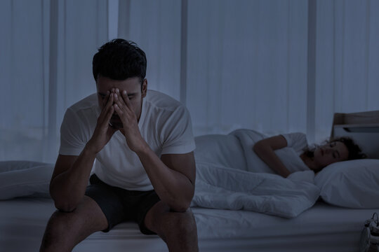 Upset depressed young man sitting on the edge of bed against his wife lying on the bed. Relationship difficulties , Problems couple.