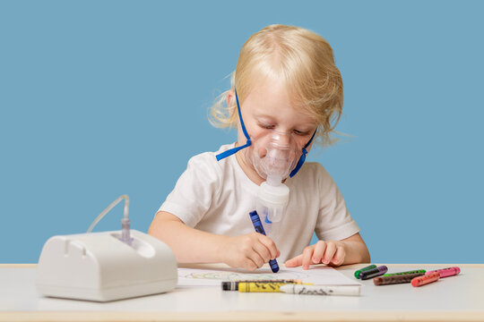 A happy child in therapeutic inhalation mask is sitting at a table and drawing with colored pencils. Treatment of colds at home. Copyspace.