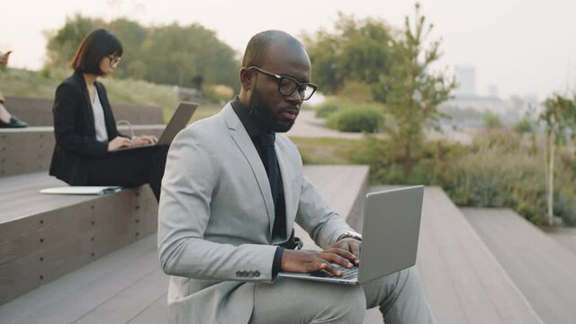 Young African American businessman in stylish formal suit sitting on stepped seating in park and working on laptop