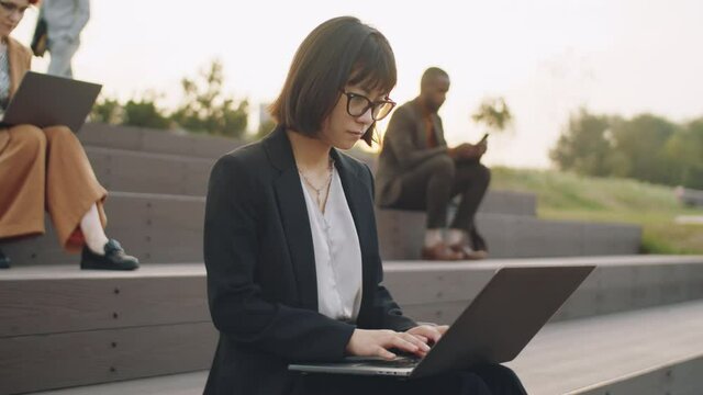 Young Asian businesswoman in formalwear and glasses sitting on stepped seating in park and typing on laptop