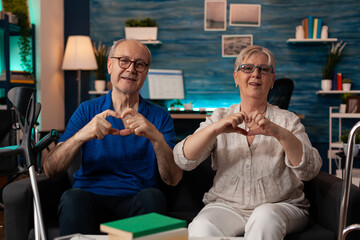 Elderly couple creating heart shape with hands while looking at camera in living room. Married people in love doing symbol of affection together and sitting on sofa at home. Senior relationship