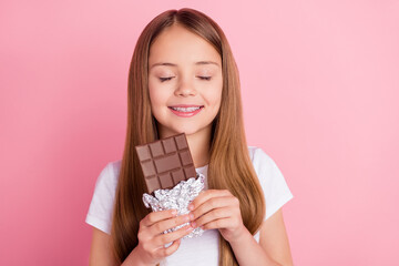 Photo of nice blond small girl eat chocolate closed eyes wear white t-shirt isolated on pink color background