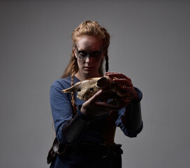 Closeup portrait of young red haired woman with black barbarian facepaint wearing medieval viking inspired costume Holding  sheep skull for ritual  posing against studio background with red lighting