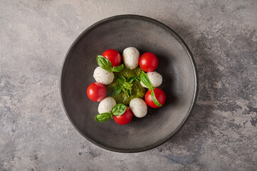 Delicious italian caprese salad with ripe tomatoes, pesto, basil and mozzarella cheese in black plate on the background as concrete. Top view 