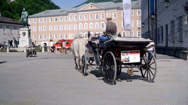 image of a horse carriage in the center of the city of Salzburg Austria where we can see the parked horse carriage and its driver from behind posing for some tourists on a sunny summer day