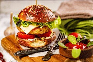 Homemade appetizing chicken burger with cheese, fresh salad and vegetables on a cutting board. Fresh lettuce leaves, tomatoes and onions. Traditional American food. White rustic background. Close-up