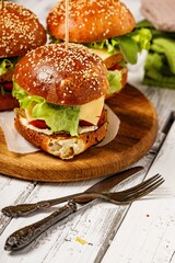 Close-up appetizing chicken burger with cheese, fresh salad and vegetables on a round cutting board. Lettuce leaves, tomatoes and onions. Traditional American fast food. Rustic background. Copy space