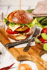 Appetizing chicken burger with cheese, fresh salad and vegetables on a cutting board. Fresh lettuce leaves, tomatoes and onions. Traditional American food. White rustic background. Vertical shot