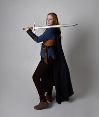 Full length, portrait of red haired woman wearing medieval viking inspired costume and flowing cloak,  Holding a long sword weapon  posing against studio background.