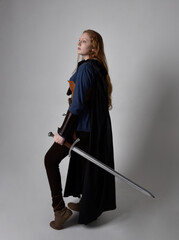 Full length, portrait of red haired woman wearing medieval viking inspired costume and flowing cloak,  Holding a long sword weapon  posing against studio background.