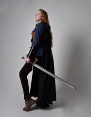 Full length, portrait of red haired woman wearing medieval viking inspired costume and flowing cloak,  Holding a long sword weapon  posing against studio background.