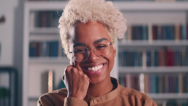 Attractive African American millennial relaxed woman smile look at camera stand indoor alone. Video call event, virtual meeting participant, happy modern cozy apartment owner portrait concept.