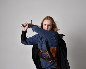 Close up portrait of red haired woman wearing medieval viking inspired costume and flowing cloak,  Holding a long sword weapon, action pose against studio background.