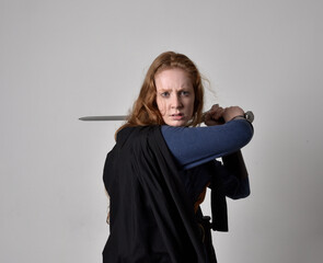 Close up portrait of red haired woman wearing medieval viking inspired costume and flowing cloak,  Holding a long sword weapon, action pose against studio background.