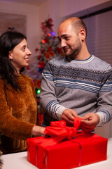 Obraz na płótnie Canvas Cheerful family opening xmas decorated gift with ribbon on it during christmas holiday standing in x-mas kitchen. Happy couple married celebrating winter season. Santa-claus tradition