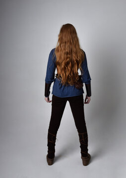 Full Length Portrait Of Red Haired Woman Wearing Medieval Viking Inspired Costume,  Standing Pose With Back To The Camera, Against Studio Background.