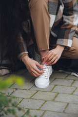 Closeup photo of young woman wearing checkered long coat, and beige pants . Lady posing on city street. Women ties laces on sneakers