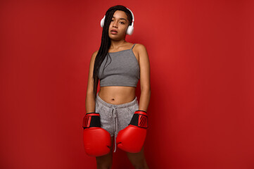 Exhausted young athlete woman boxer, sportswoman wearing wireless headphones, looks at camera posing with red boxing gloves against a red background with copy space for ad. Contact martial art concept